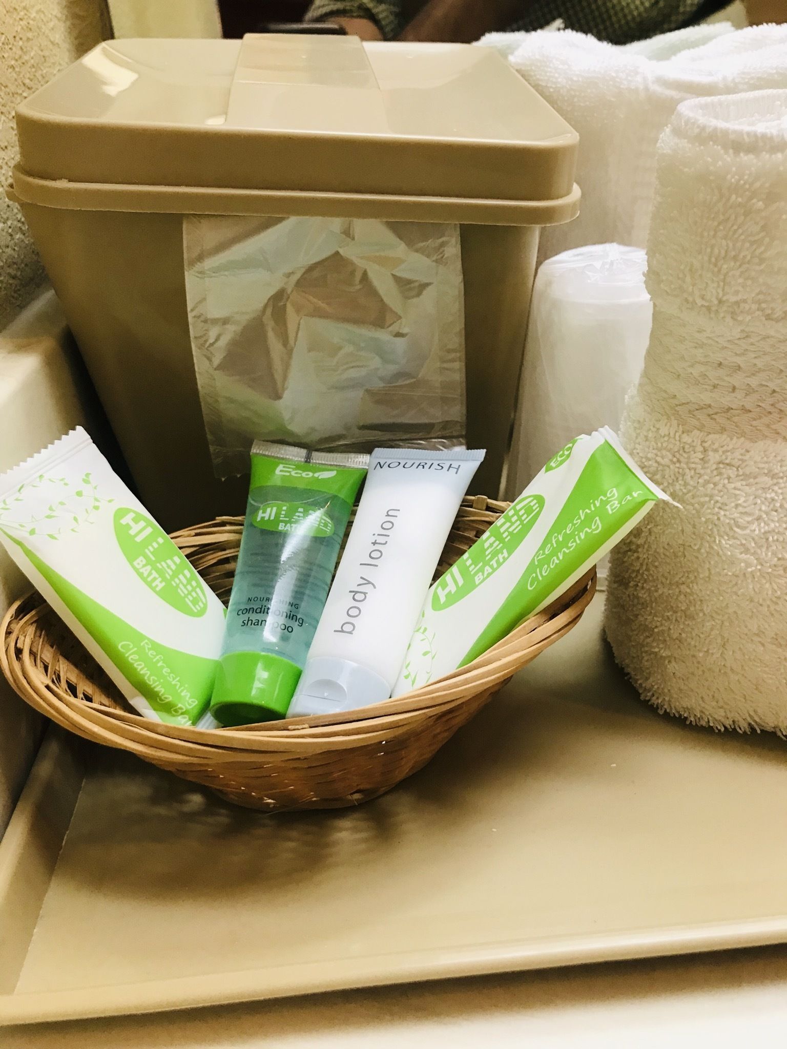 A basket containing toiletries including a body lotion tube, a green bath product, and a green cleansing bar, placed on a tray with a beige container and white towels in the background.