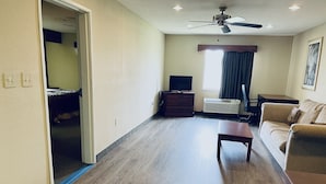 A spacious living room with a wooden floor, a ceiling fan, a television on a wooden dresser, a sofa, and a window with curtains. - America's Best Inn and Suites Flowood (Flowood)