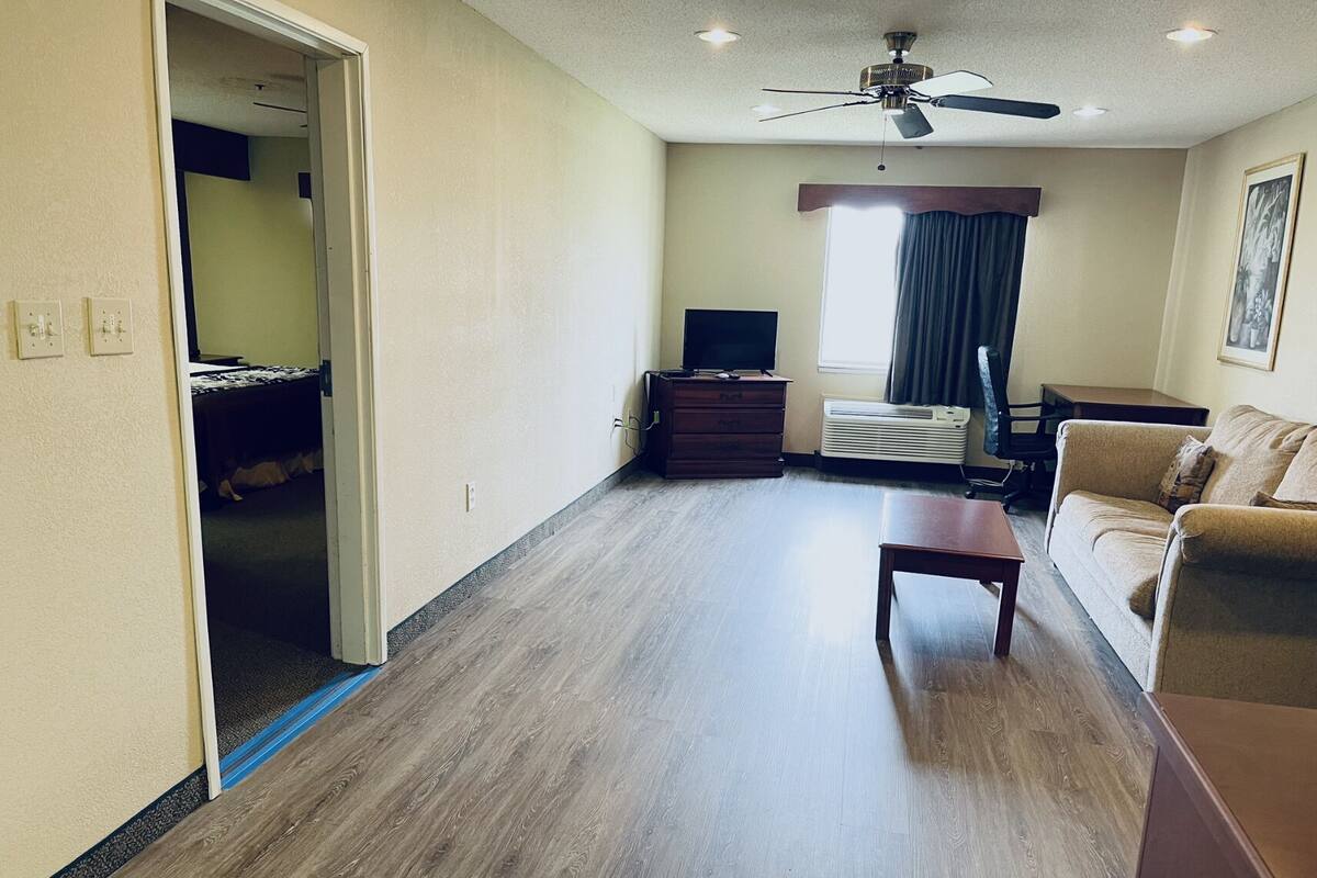 A spacious living room with a wooden floor, a ceiling fan, a television on a wooden dresser, a sofa, and a window with curtains.
