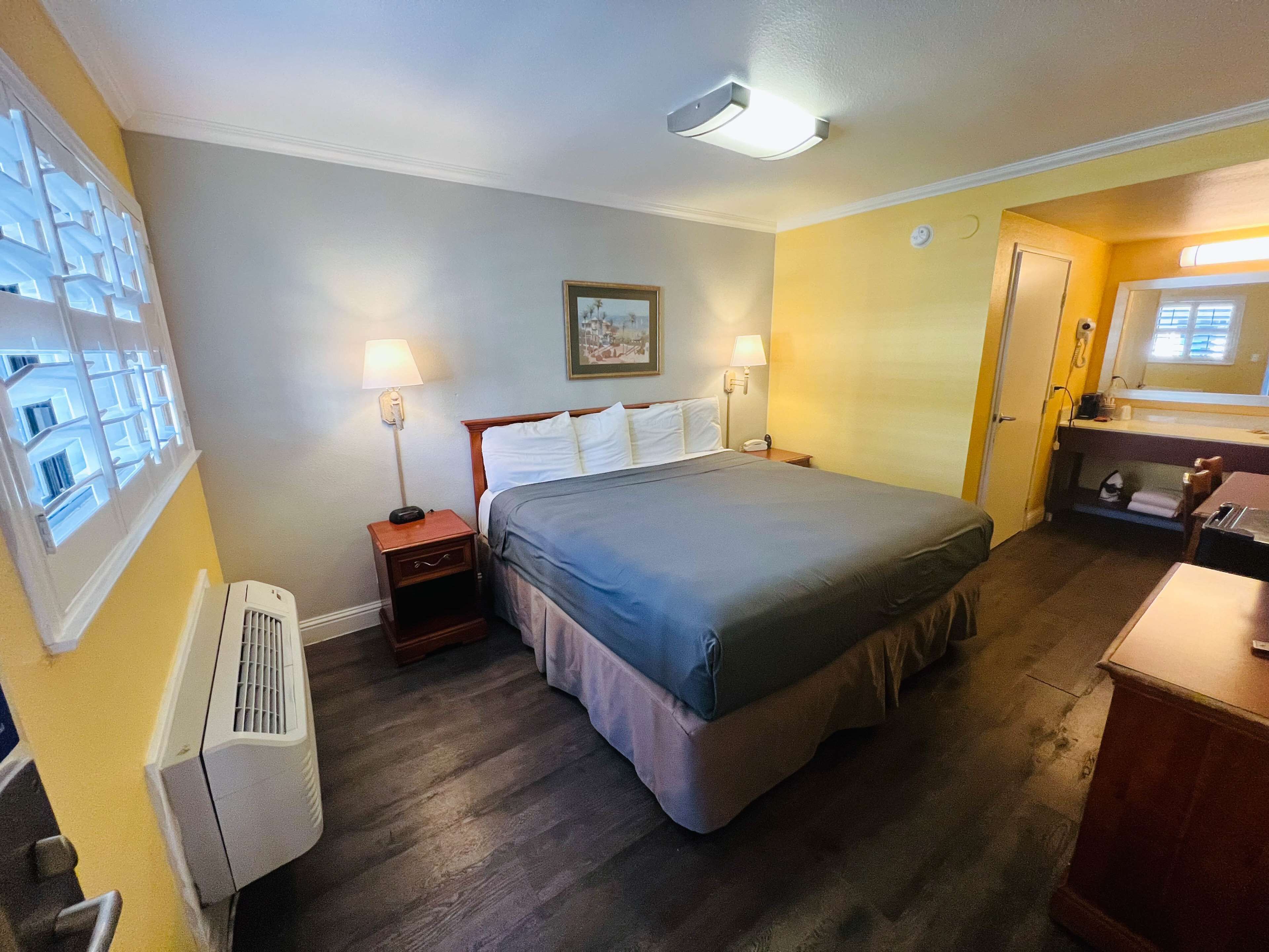 A hotel room with a bed, bedside lamps, a nightstand, a desk, a chair, a window with blinds, and a wall-mounted air conditioner.