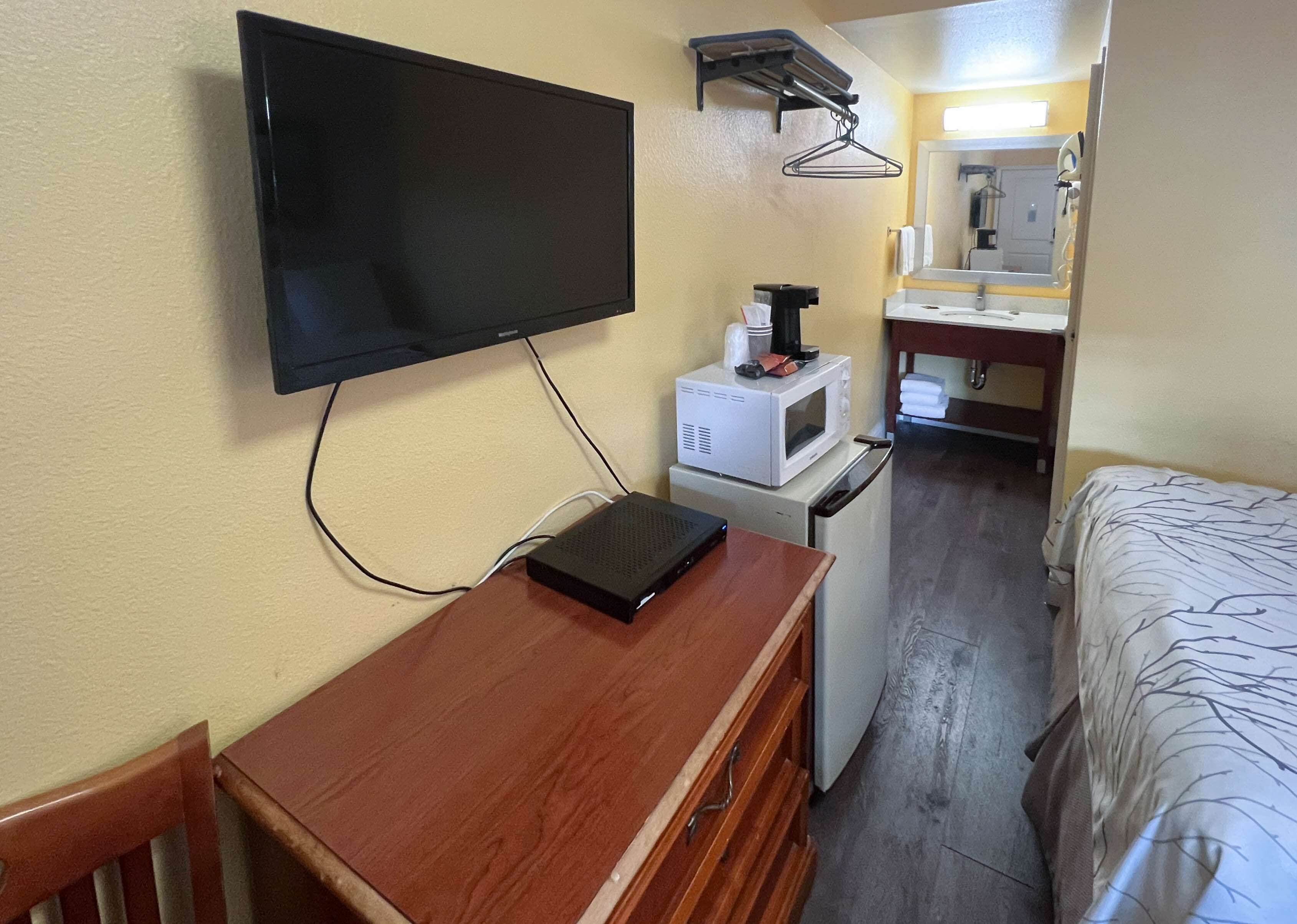 A compact hotel room with a flat-screen TV mounted on the wall, a small kitchenette with a microwave, and a bed with a patterned bedspread.