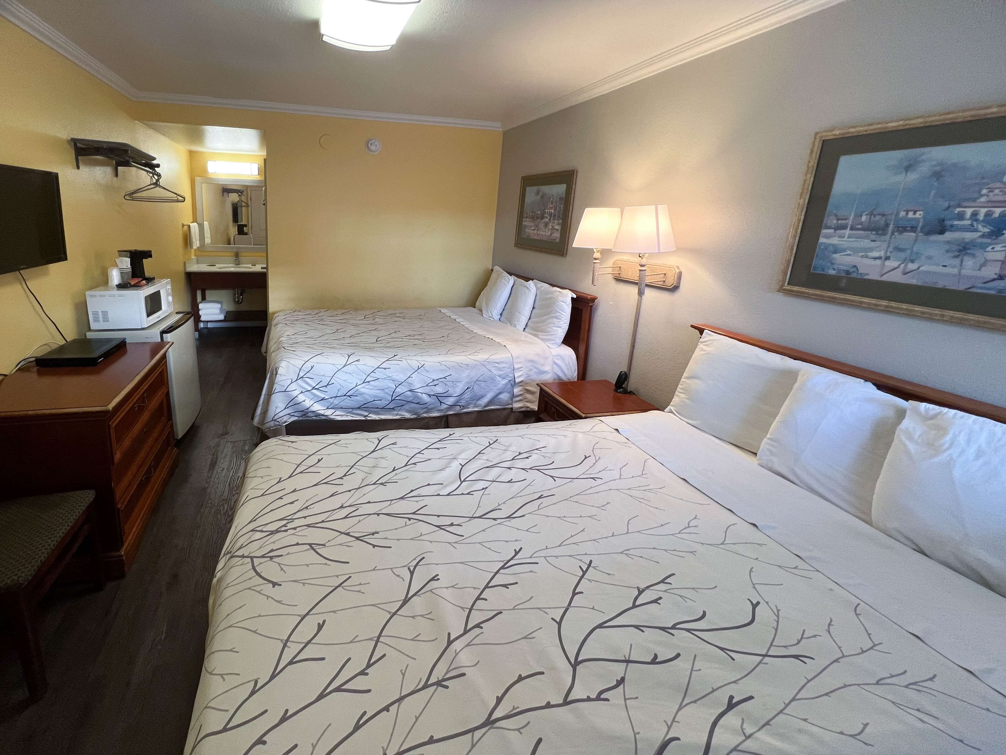 A hotel room with two beds, a desk, a microwave, a lamp, and a painting on the wall.