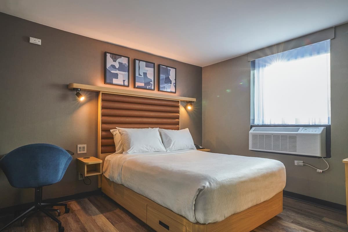 A hotel room with a bed, a desk, a chair, a window with an air conditioner, and framed artwork on the wall.