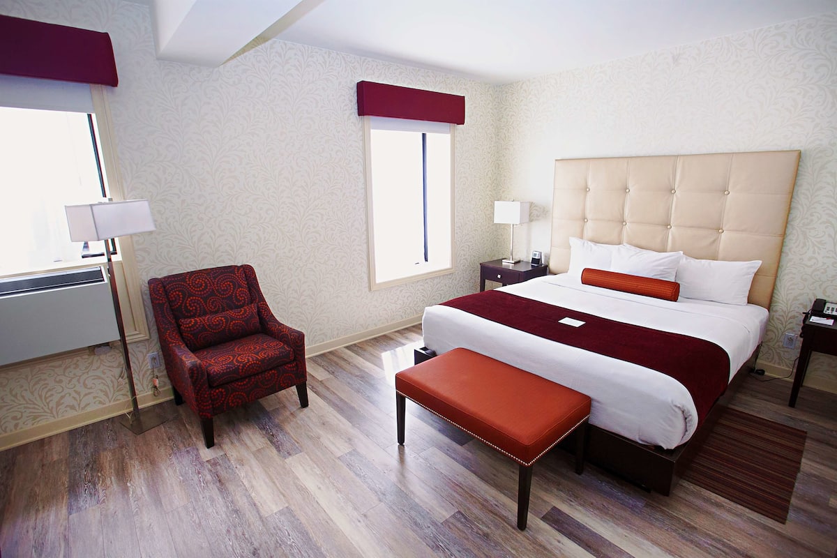 A hotel room with a large bed, a red armchair, a bench, and a window with a view.