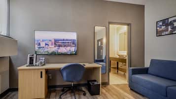 A hotel room with a desk, chair, sofa, television, mirror, and a view of the cityscape.