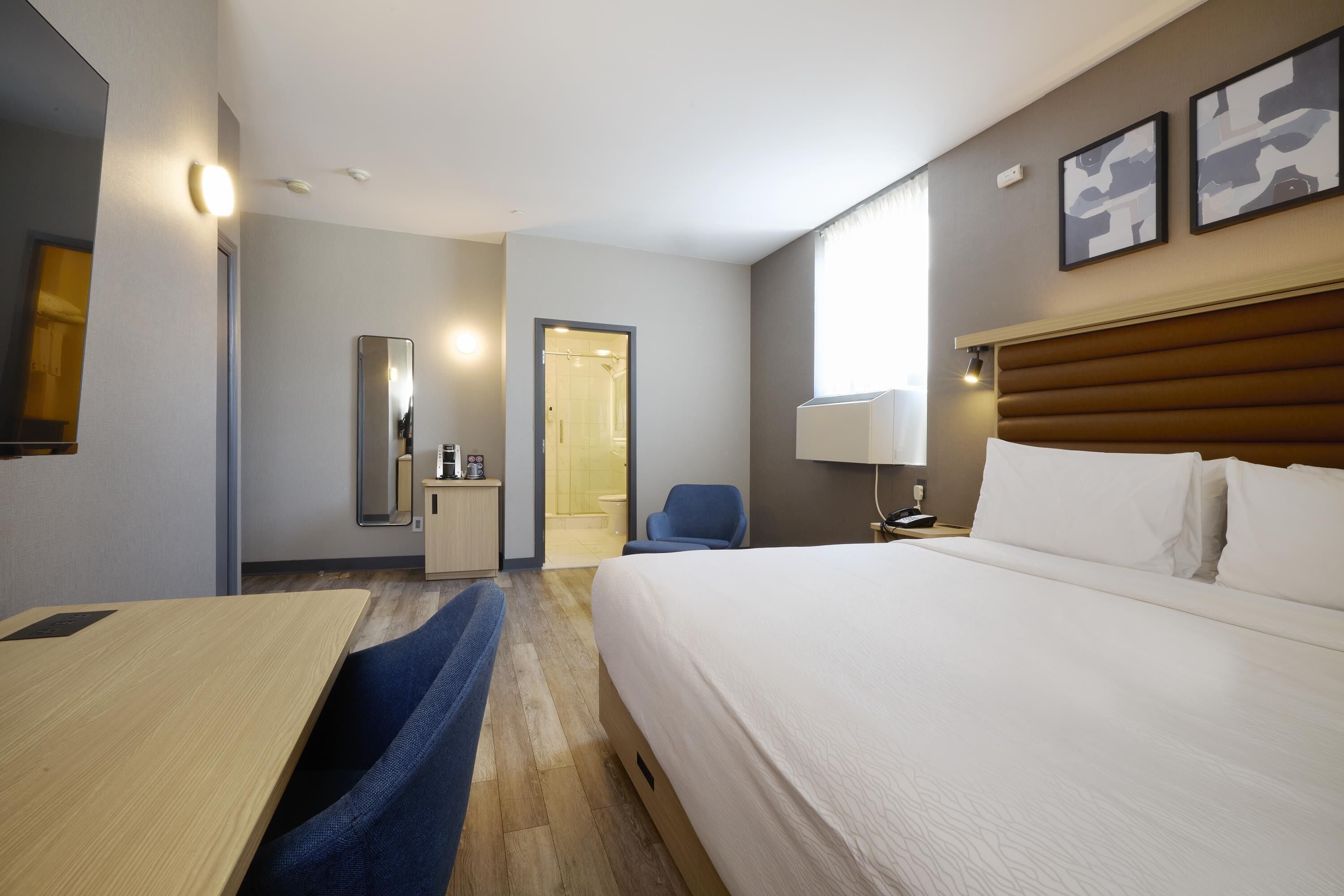 Quarto de hotel com cama, escrivaninha, cadeira, espelho, porta de acesso a um banheiro e uma luminária na parede.