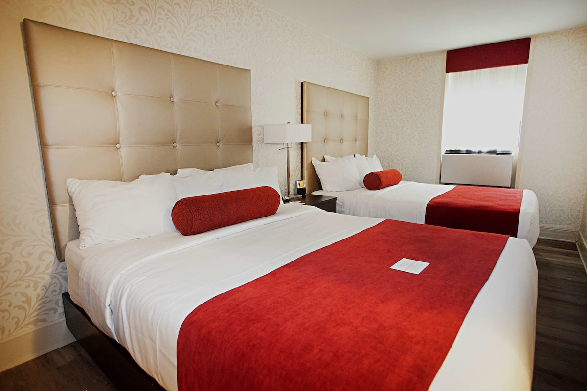 A hotel room with two beds, each with a red accent pillow, a nightstand with a lamp, and a window with a red curtain.
