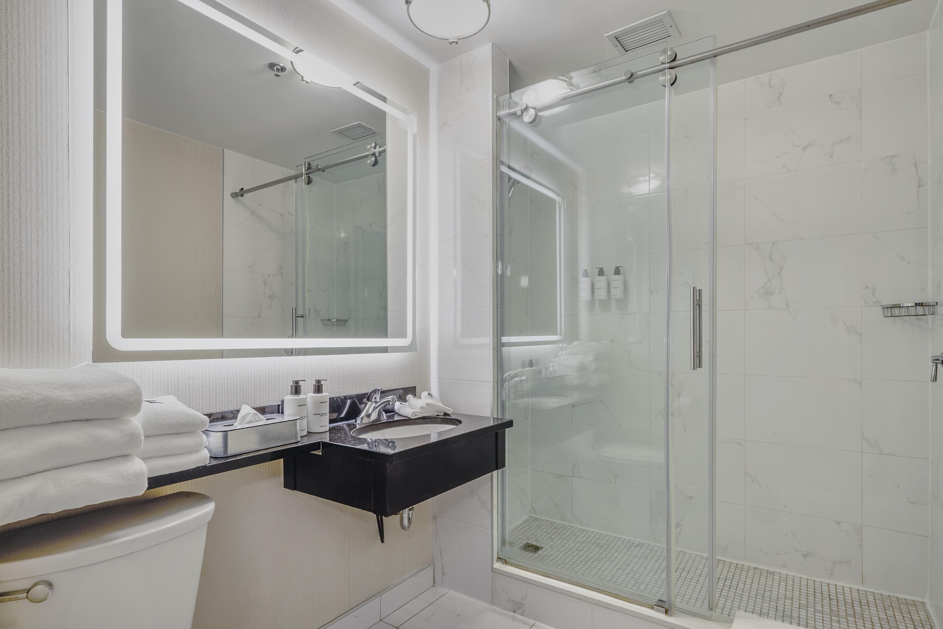 Une salle de bain moderne avec une douche à parois de verre, un grand miroir et un plan de toilette noir sur lequel sont posés des articles de toilette.