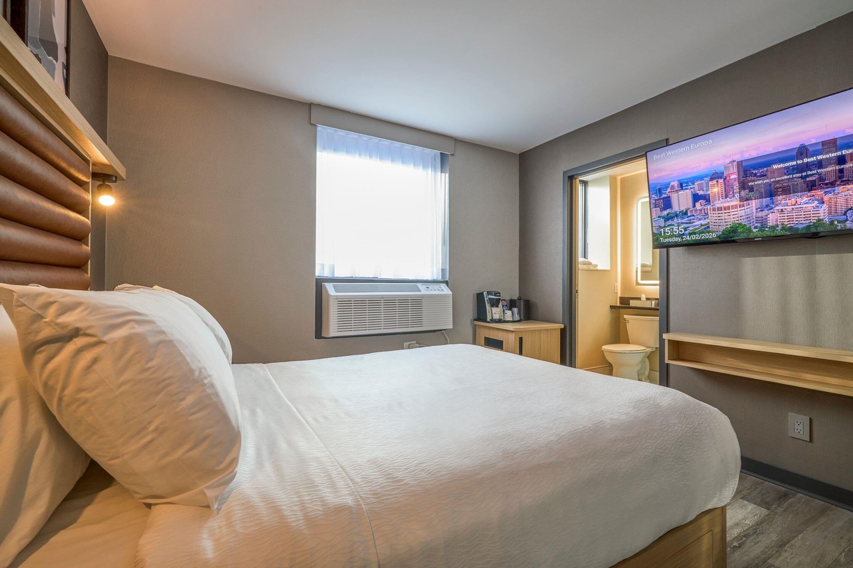 A hotel room with a bed, an air conditioning unit, a television, a window with curtains, and a small table with a coffee maker.