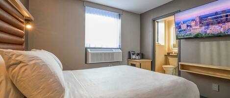 A hotel room with a bed, an air conditioning unit, a television, a window with curtains, and a small table with a coffee maker.