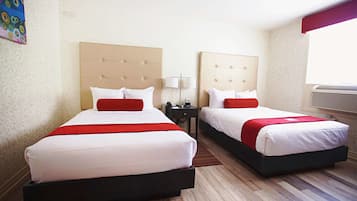 Une chambre d’hôtel avec deux lits, chacun recouvert d’une courtepointe rouge et blanche, une petite table de chevet avec une lampe, et une grande fenêtre avec des stores.