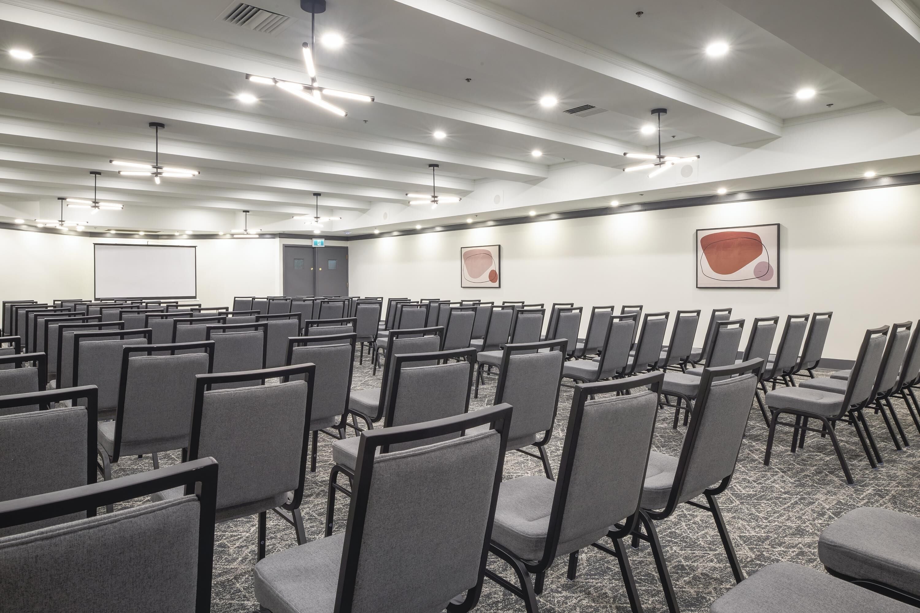 A conference room with rows of chairs, a projector screen, and two abstract paintings on the walls.