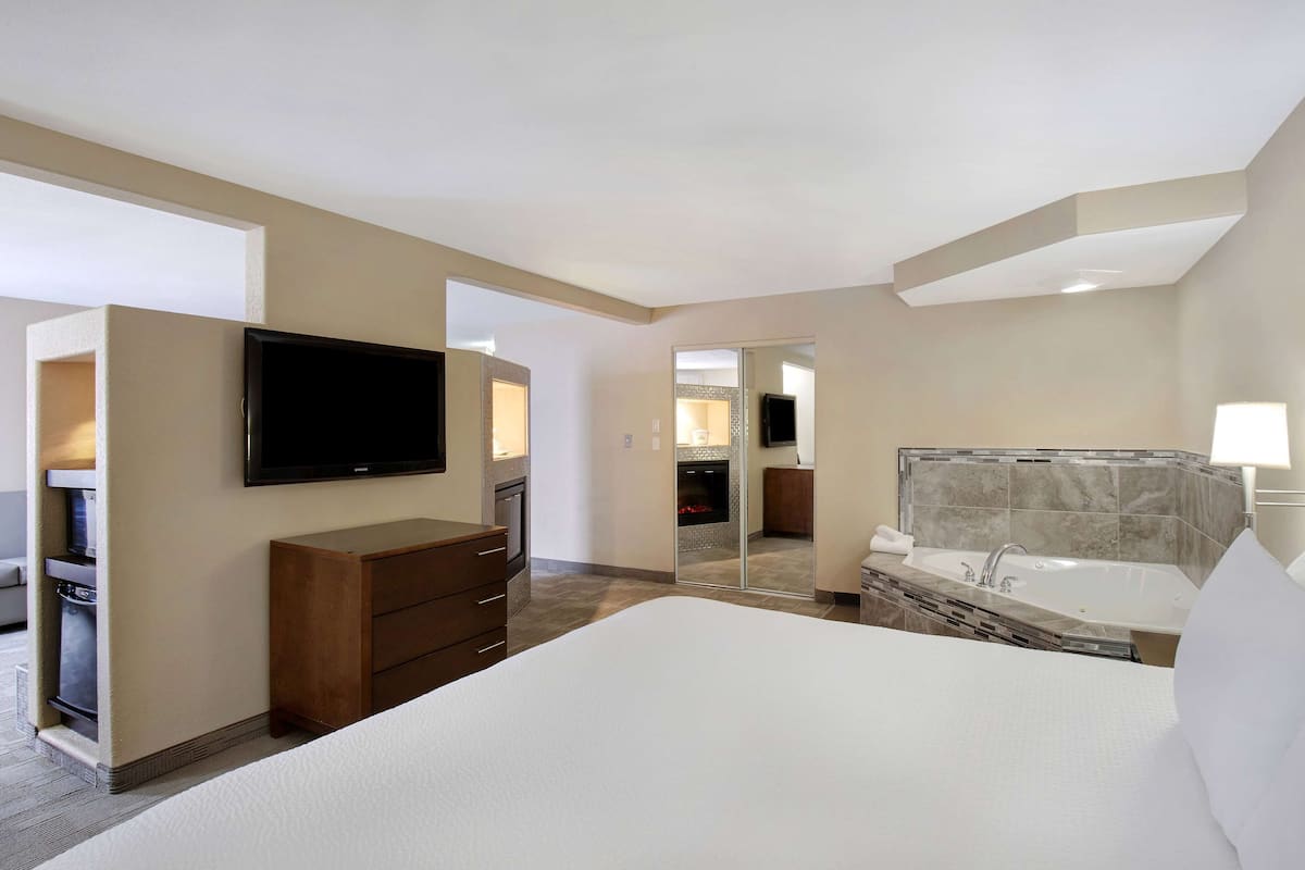 A hotel room with a large bed, a TV mounted on the wall, a wooden dresser, a bathroom with a bathtub, and a mirror.