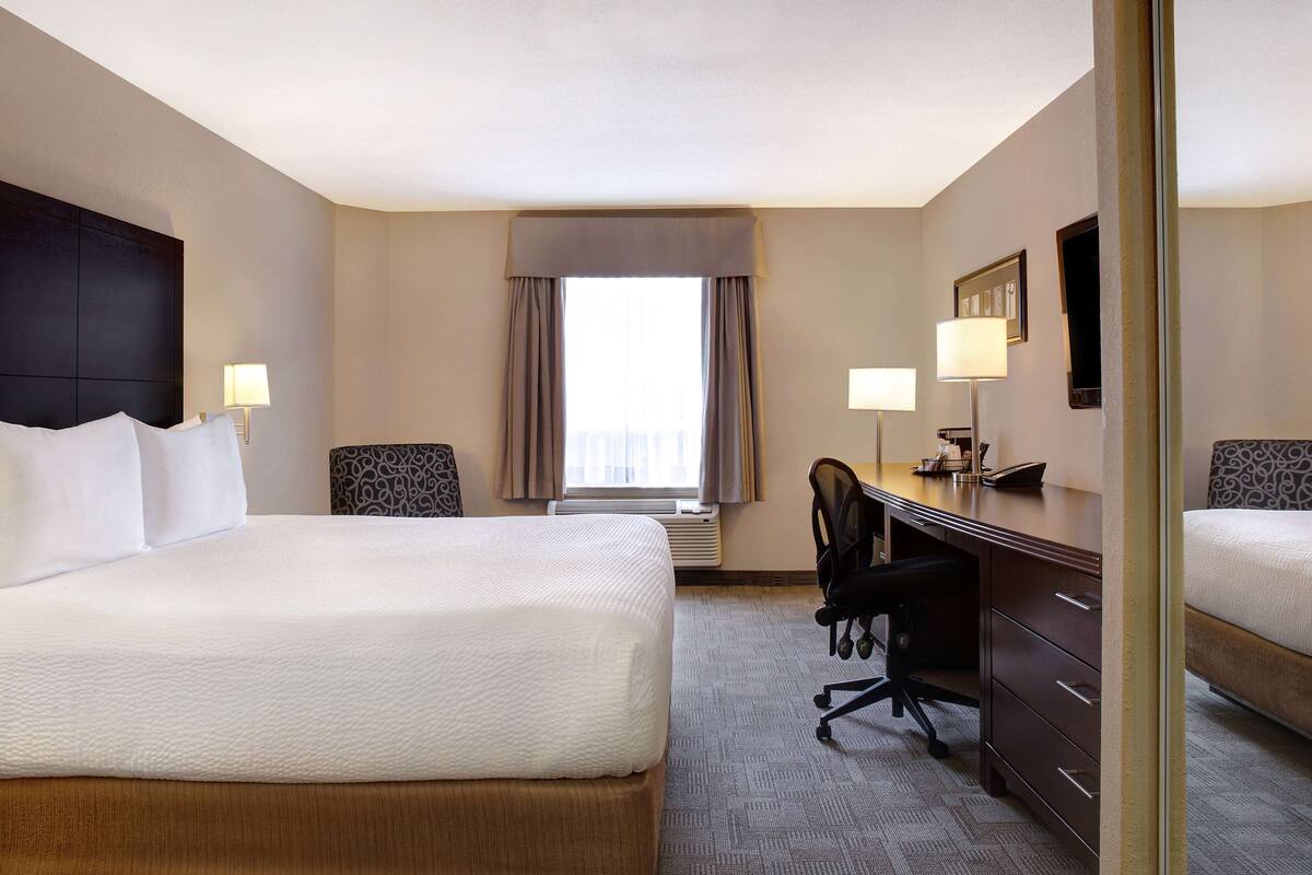 A hotel room with a large bed, a desk with a chair, a lamp, and a window with curtains.