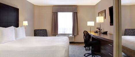 A hotel room with a large bed, a desk with a chair, a lamp, and a window with curtains.