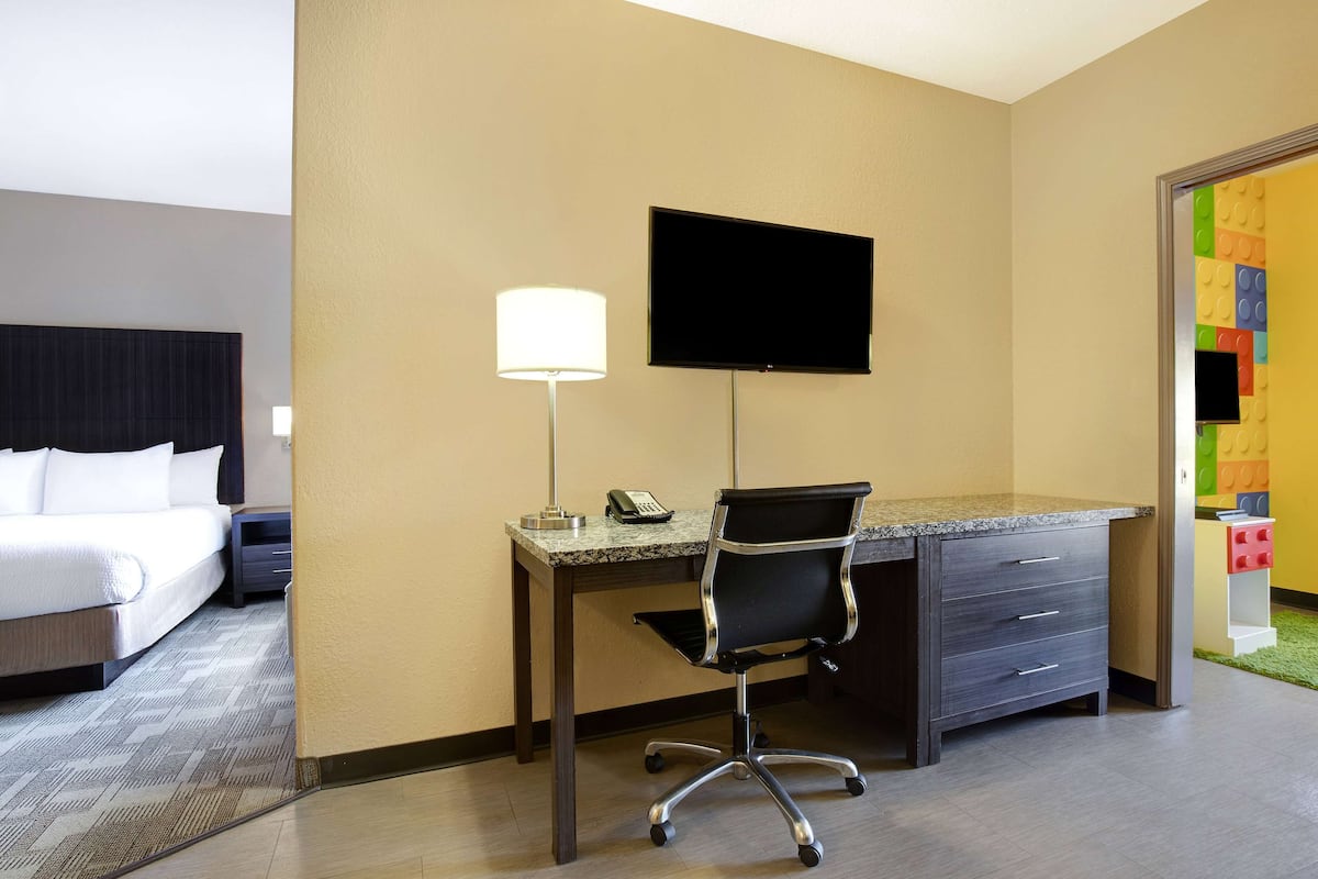 A hotel room with a bed, desk, chair, lamp, TV, and a colorful wall decoration.