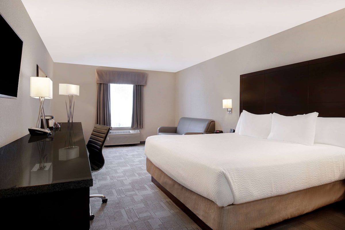 A hotel room with a large bed, a desk with a chair, a flat-screen TV, and a window with curtains.