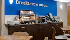 A breakfast area with a blue wall, a self-service station, and wooden tables and chairs.