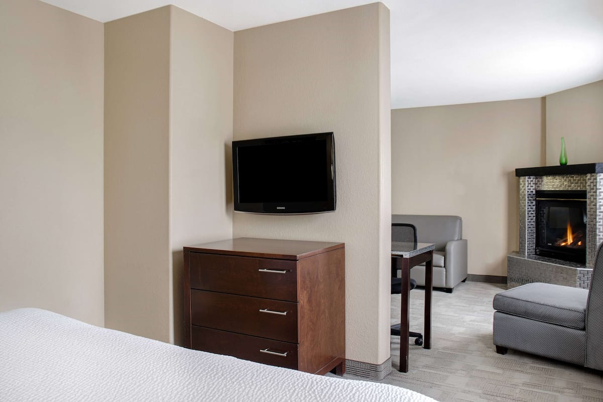 A hotel room with a bed, a wooden dresser, a flat-screen TV mounted on the wall, a gray sofa, and a fireplace.