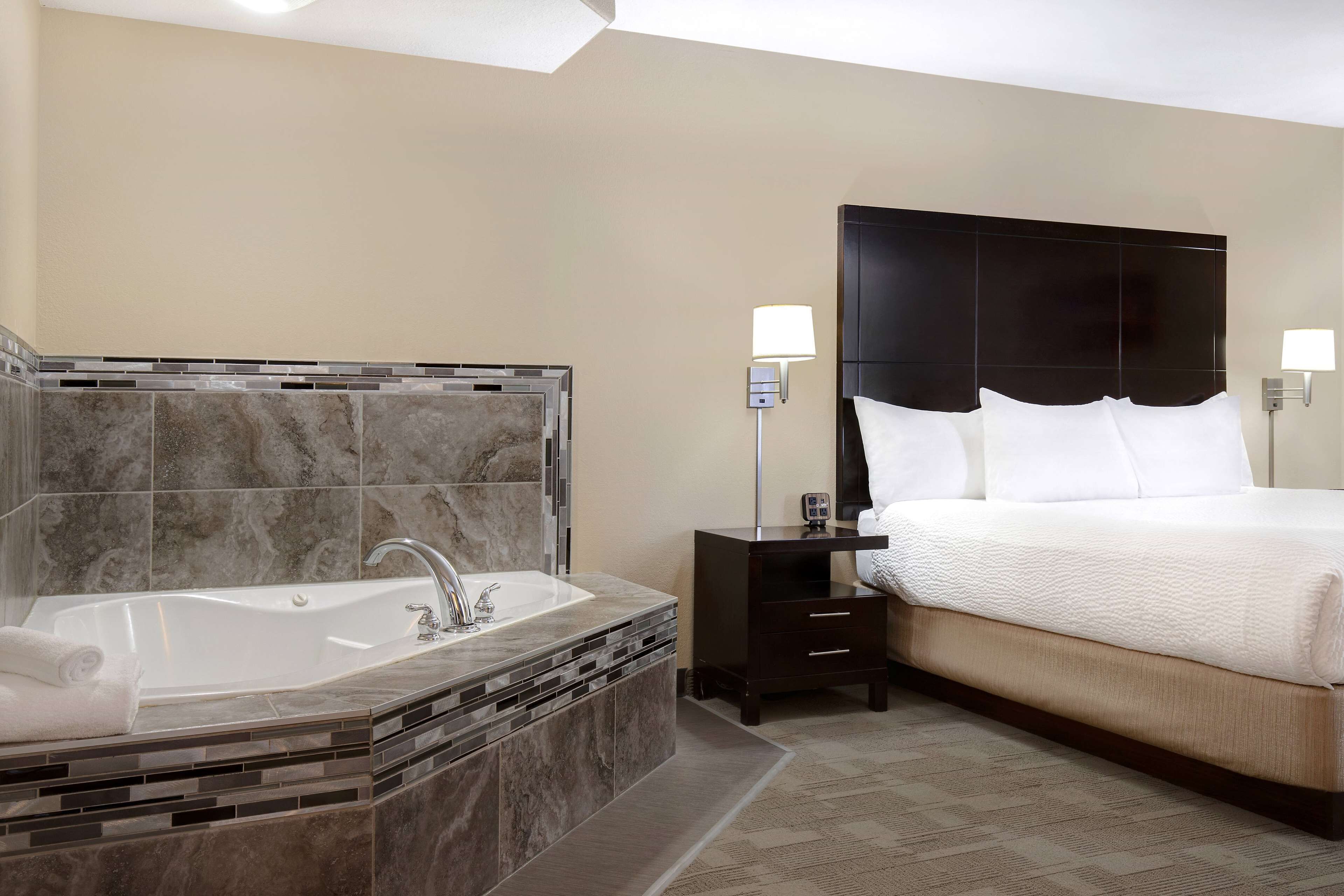 Een moderne hotelkamer met een groot bad, een bed met witte lakens en een nachtkastje met een lamp.