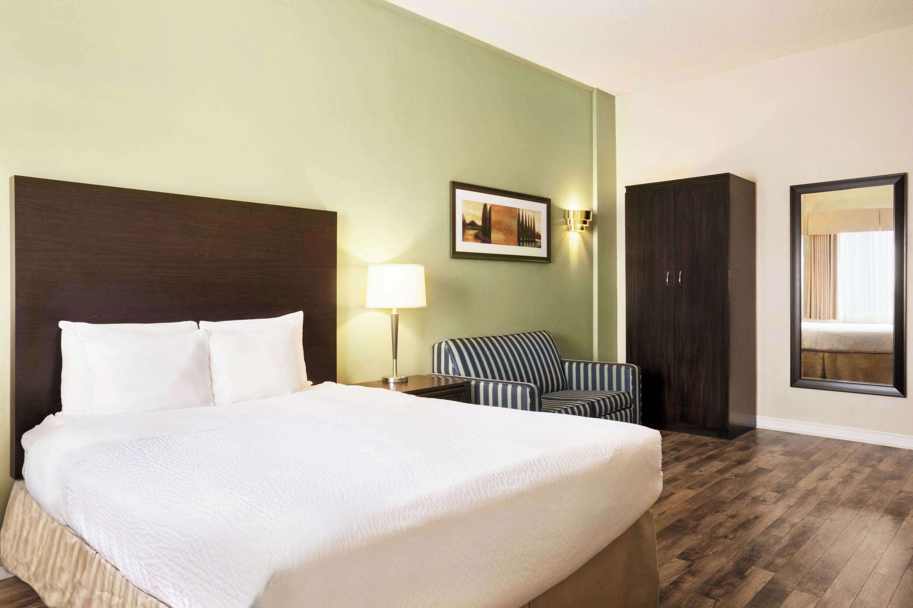 A hotel room with a large bed, a bedside lamp, a striped armchair, a dark wooden wardrobe, and a framed picture on the wall.