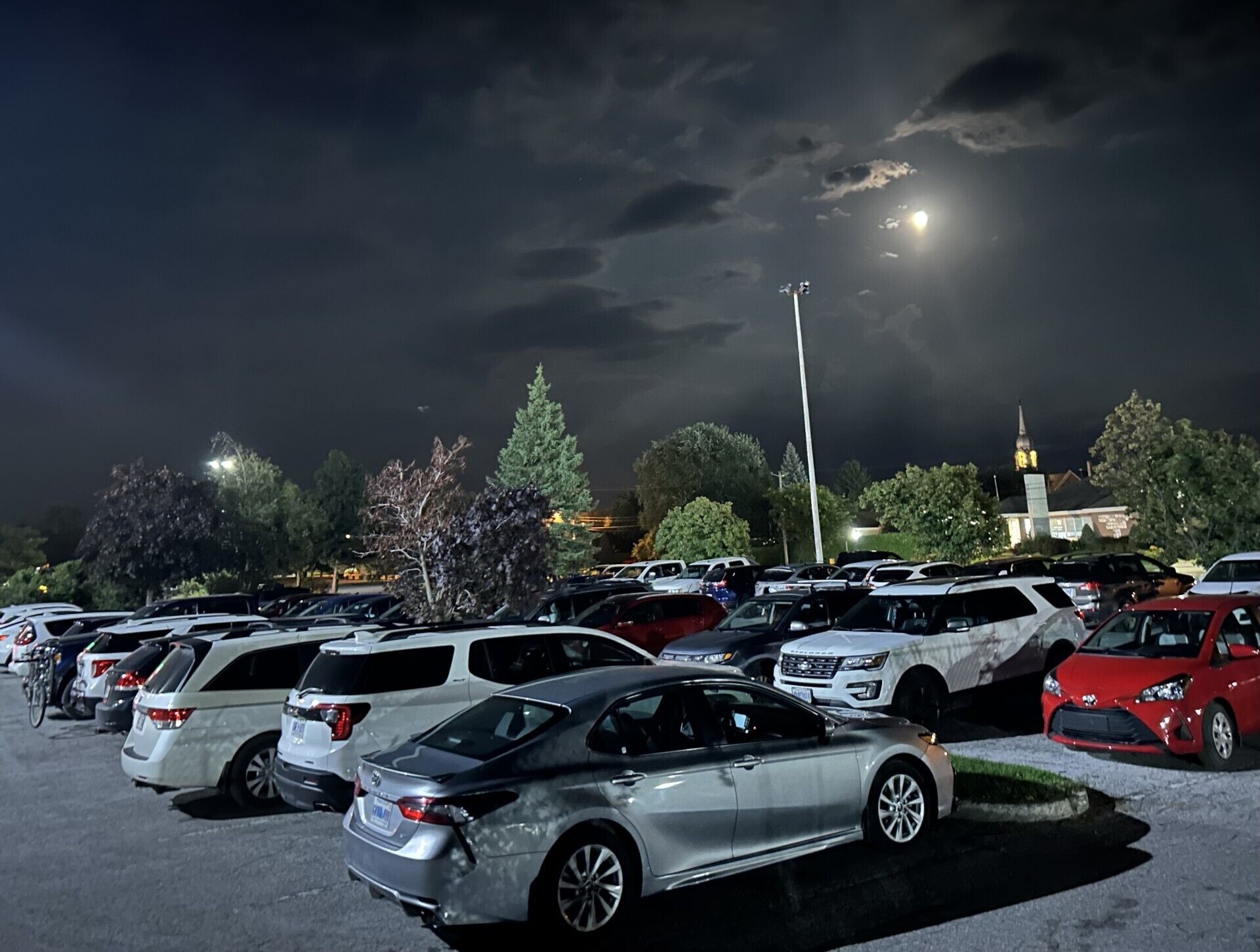Un estacionamiento de noche con varios autos y un edificio con una torre al fondo.