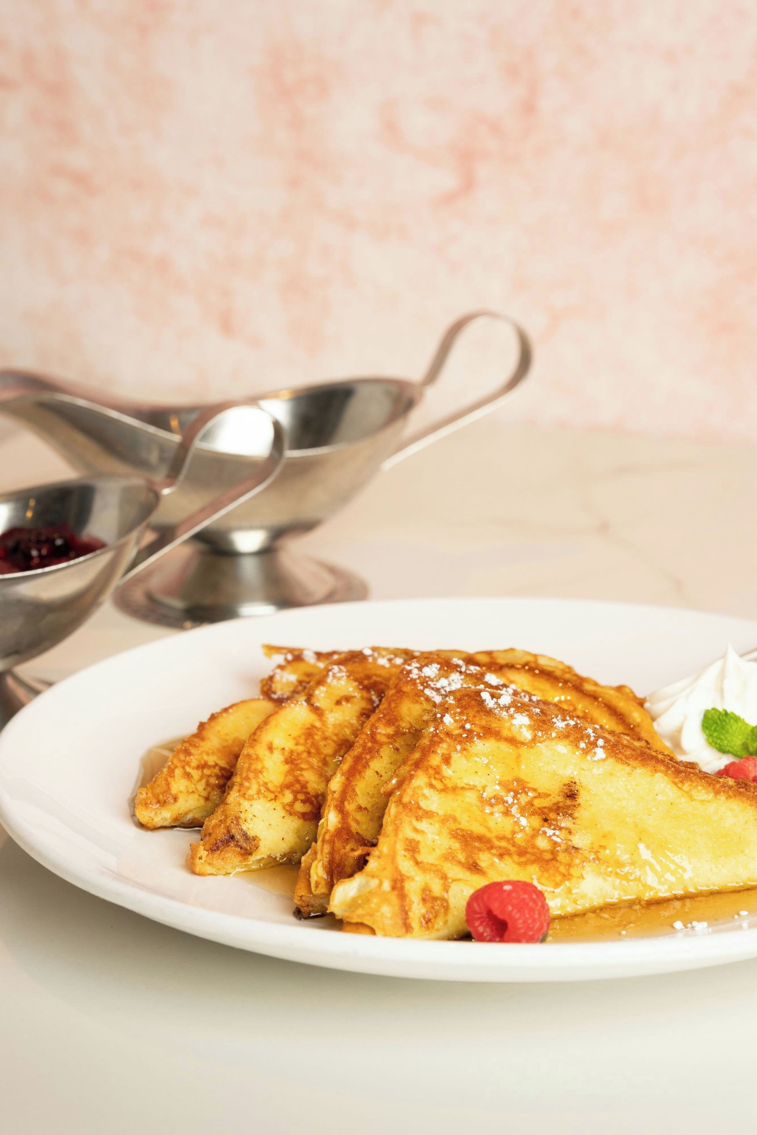 A plate of crepes with syrup and powdered sugar, a raspberry, and whipped cream on a white plate.