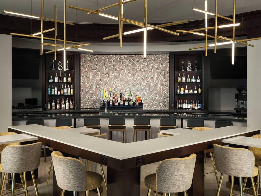 Uma área de bar com balcão central, diversas opções de assento e parede decorativa com padrão em mosaico.