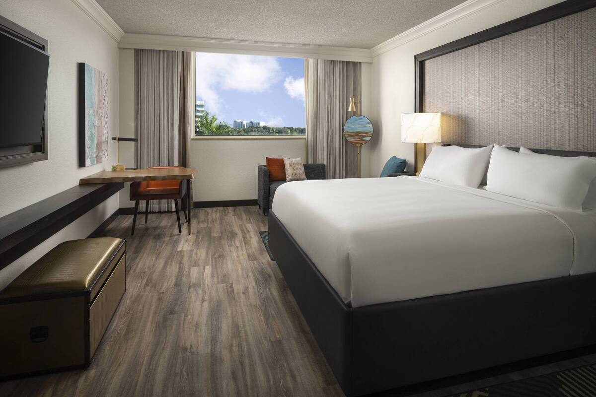 A hotel room with a large bed, a desk, a chair, a small table, a lamp, and a suitcase.