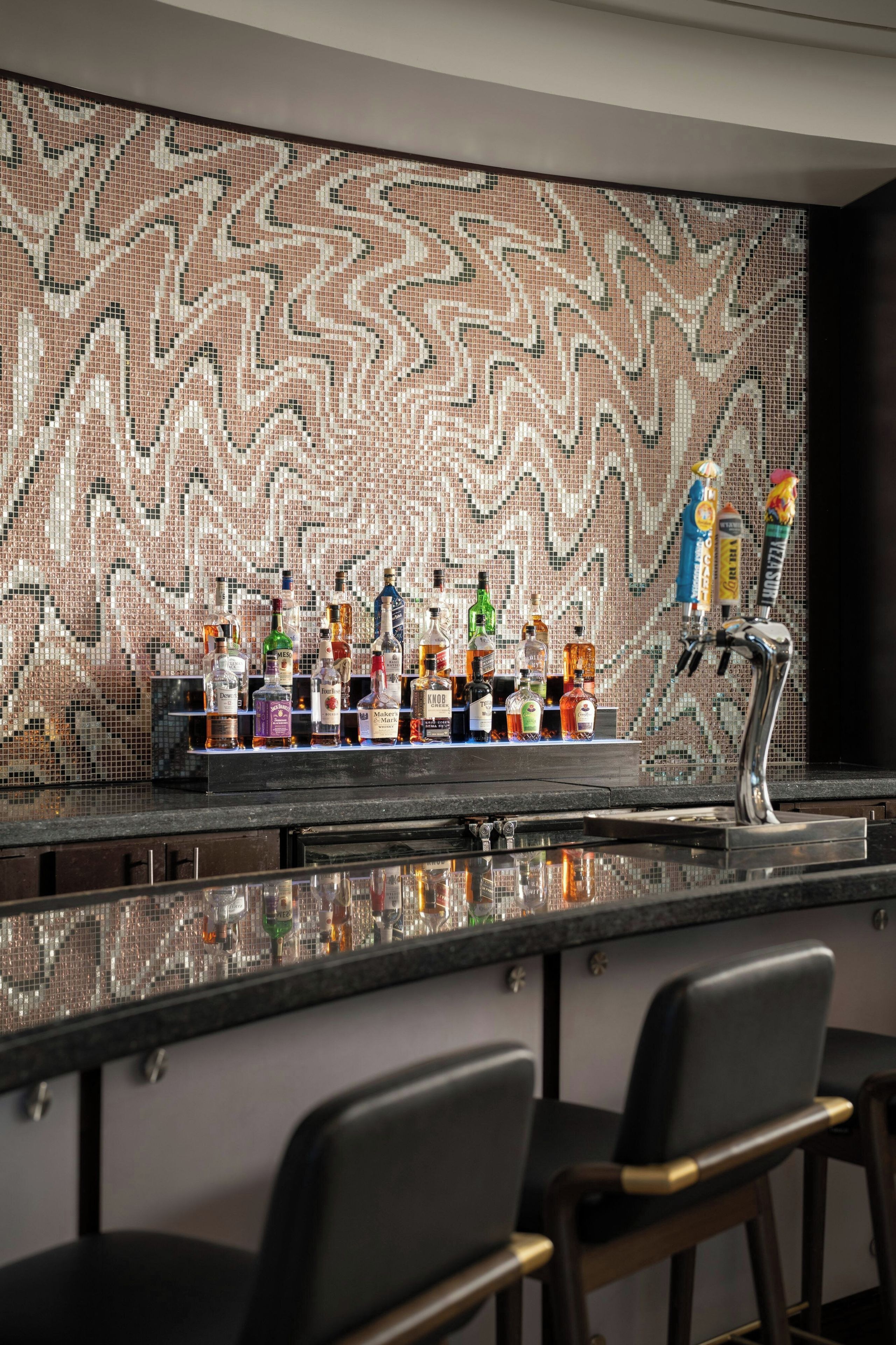 A bar area with a mosaic tile wall, a variety of liquor bottles, and two bar stools.