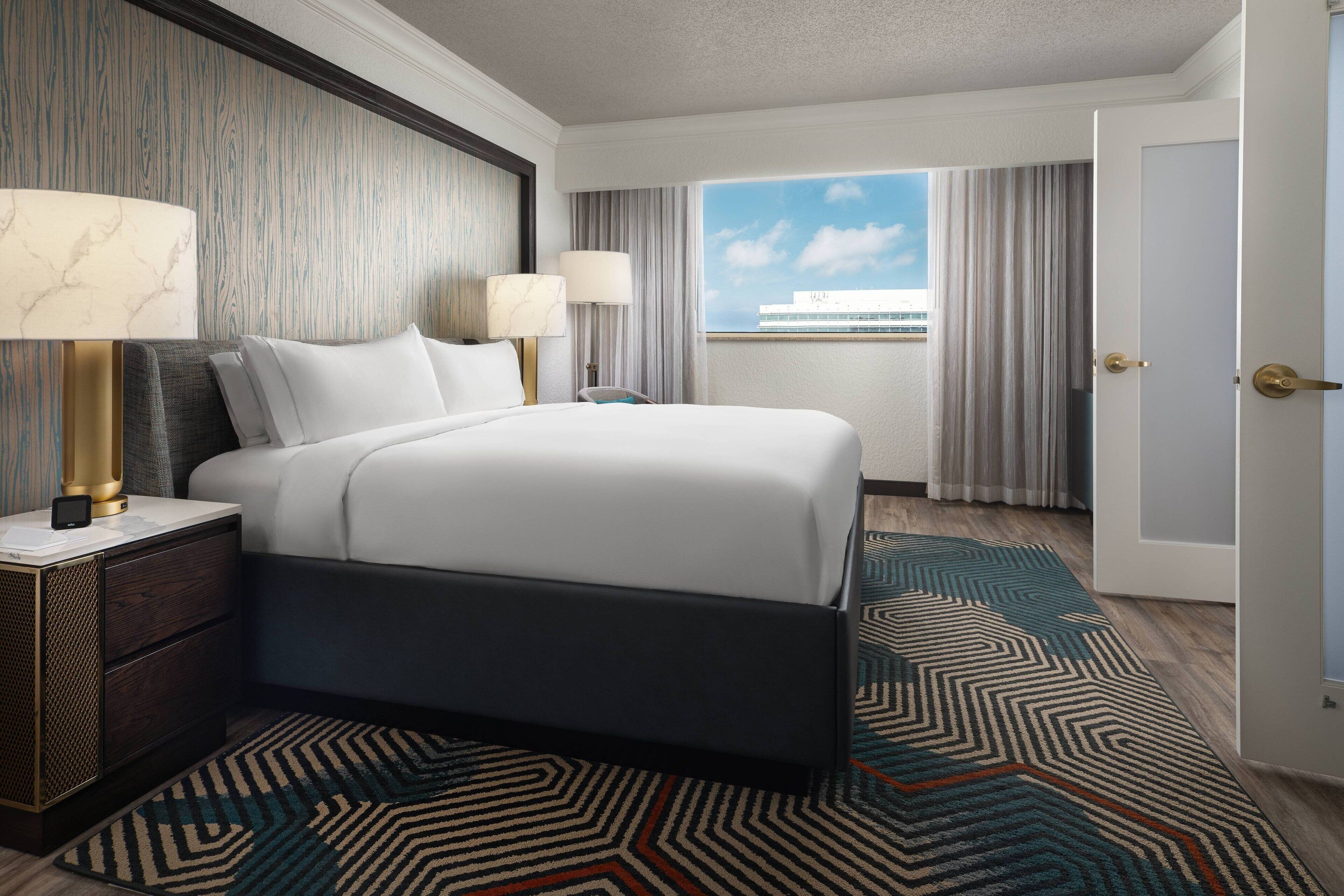 A hotel room with a large bed, two bedside tables with lamps, a patterned rug, and a view of a building through the window.