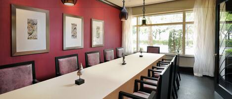 A conference room with a long table, chairs, framed artwork on the wall, and large windows with curtains.