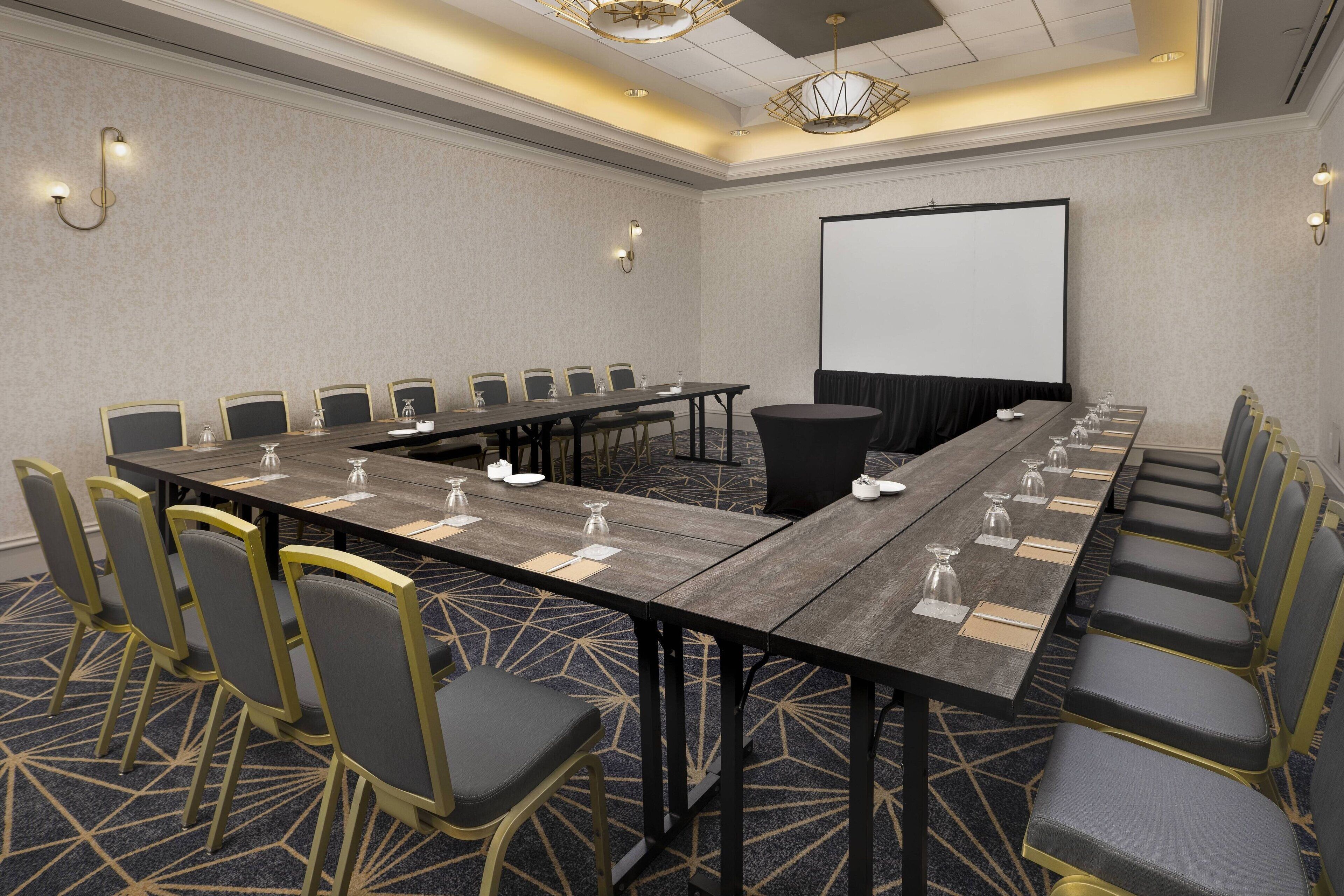 A conference room with a long table, multiple chairs, a projector screen, and a carpeted floor.