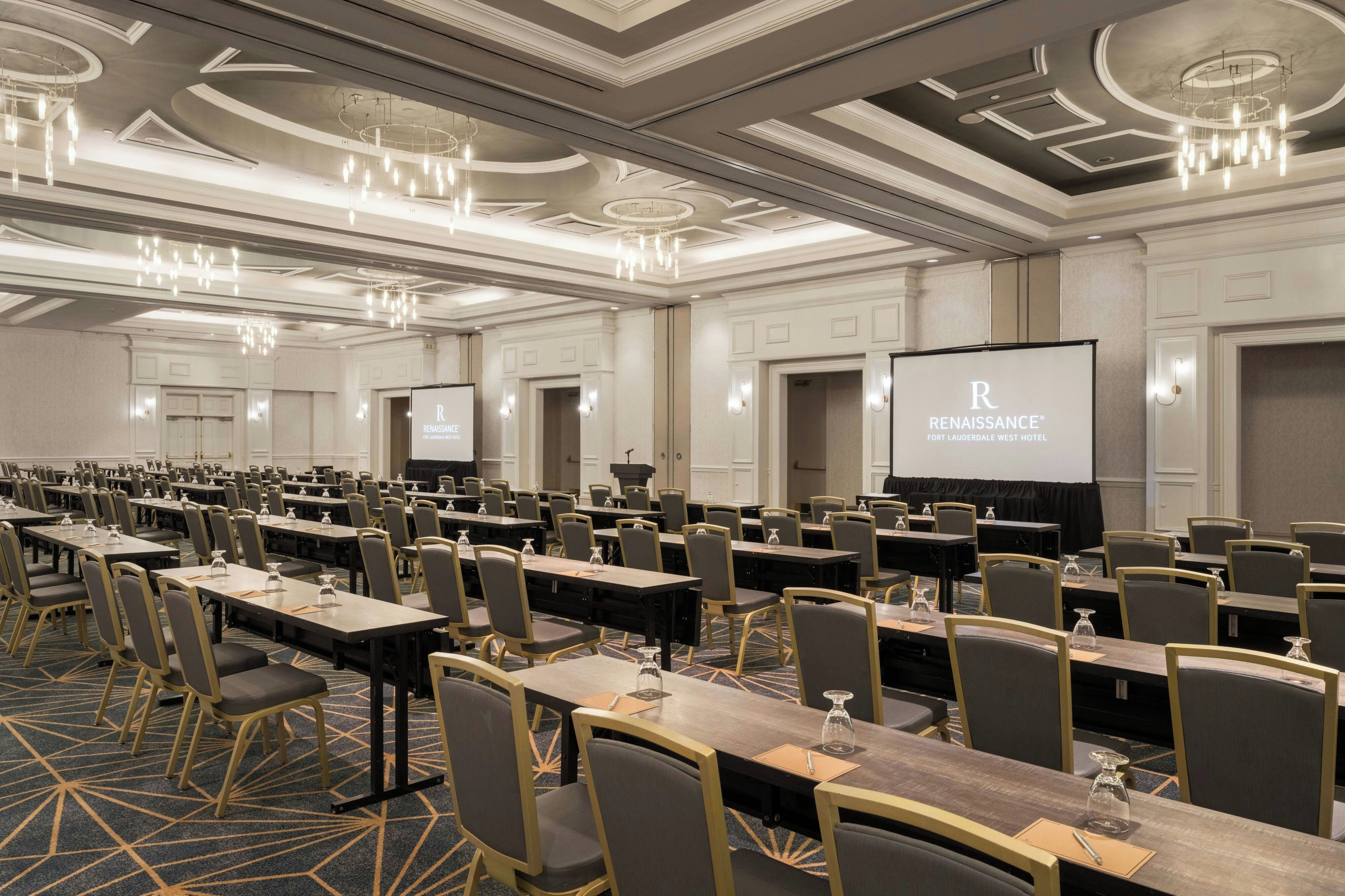A conference room with rows of tables and chairs, a large screen, and a speaker at the front.