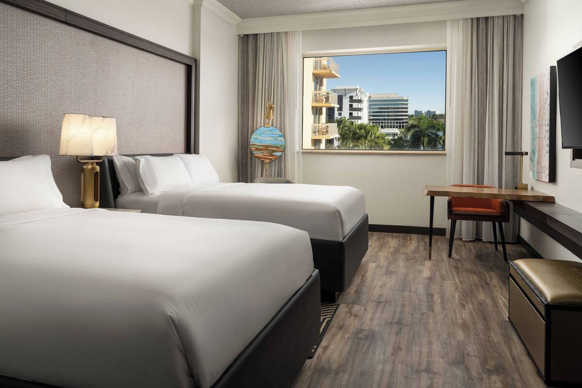 A hotel room with two beds, a desk, a chair, a TV, and a window with a city view.