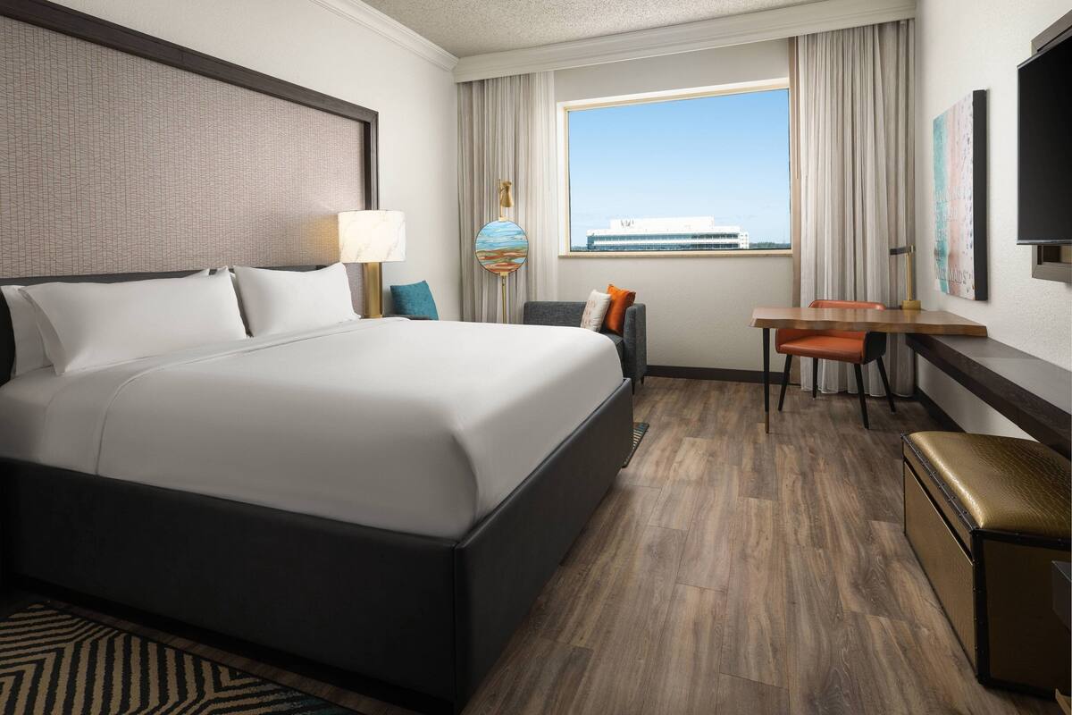A hotel room with a large bed, a desk, a chair, a television, and a window with a view of a building.