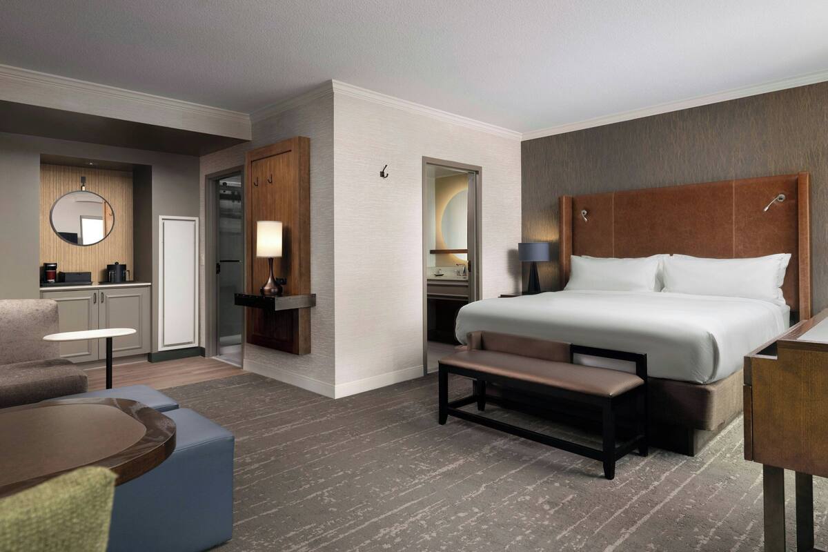 A hotel room with a large bed, a desk, a chair, a small table, and a bench.