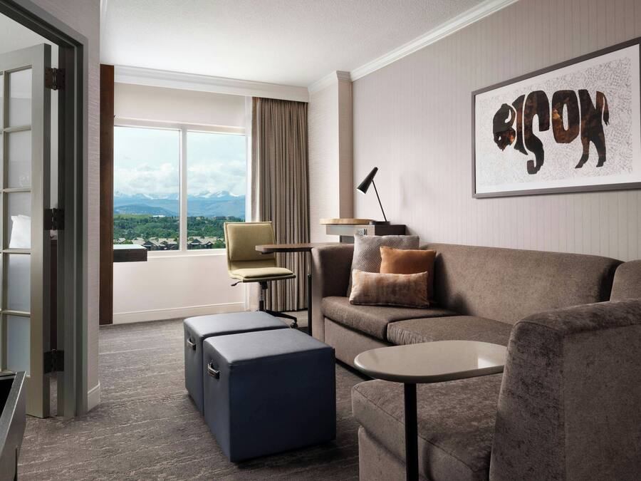 Ein Hotelzimmer mit Sofa, Hocker und einem kleinen Tisch. Ein großes Fenster bietet Blick auf Berge.