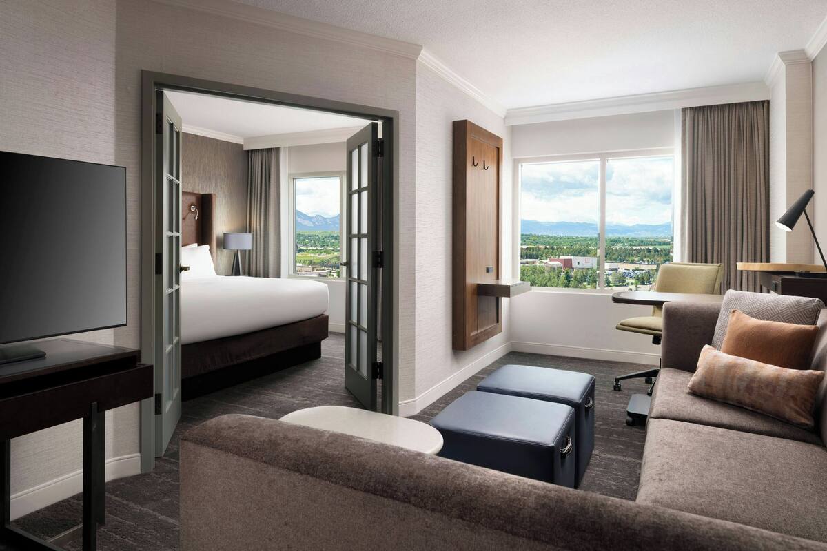 A hotel room with a bed, a sofa, a desk, and a TV. There is a large window with a view of mountains and a city.