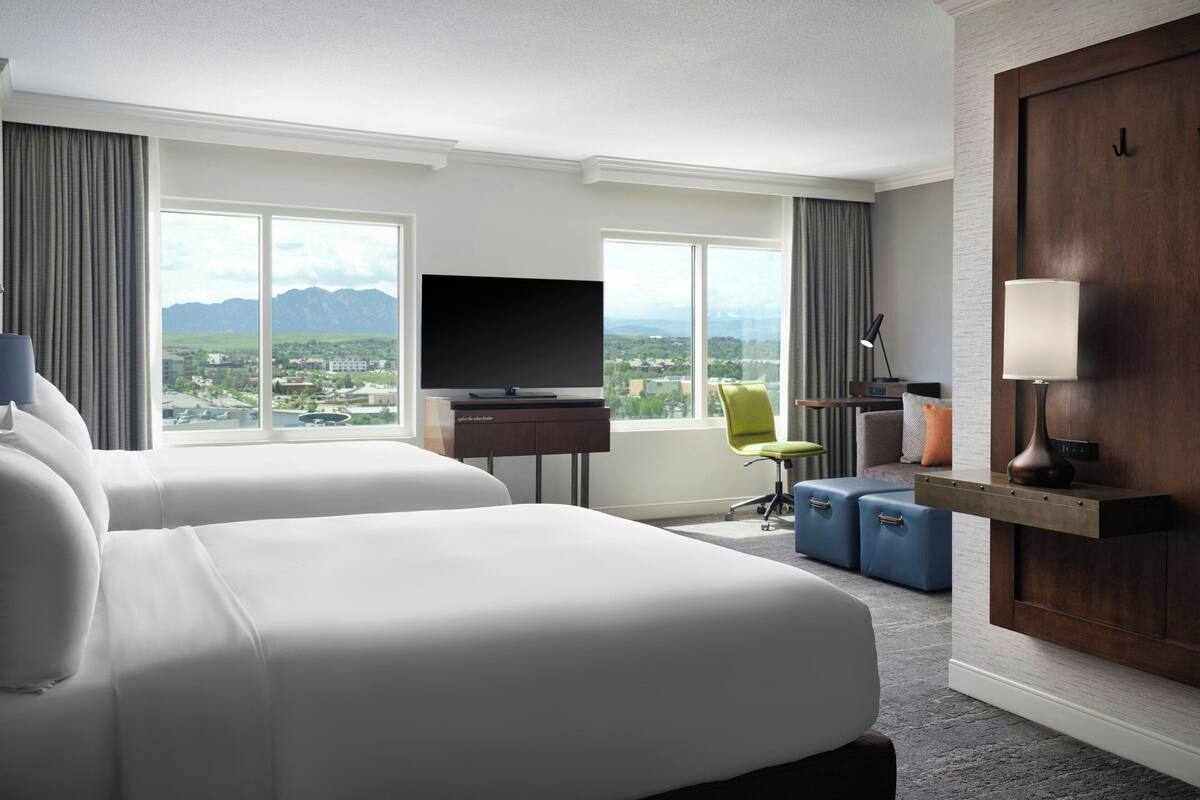 A hotel room with two beds, a TV, a desk with a lamp, a chair, and a view of mountains.
