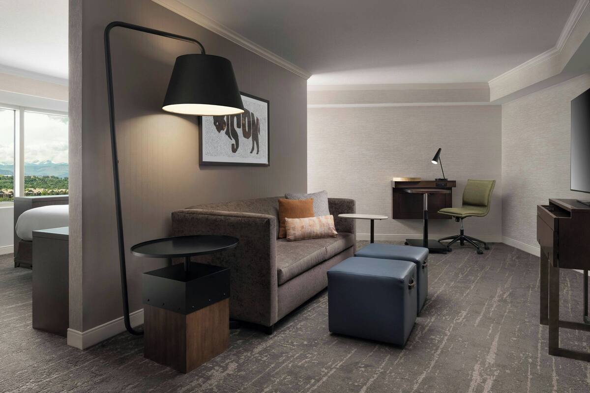 A modern hotel room with a sofa, ottoman, side table, lamp, and a desk with a chair.