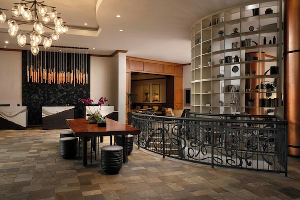 A modern lobby with a wooden reception desk, a seating area with a curved bench, a decorative light fixture, and a metal railing. - Renaissance Boulder FlatIron Hotel (Broomfield)
