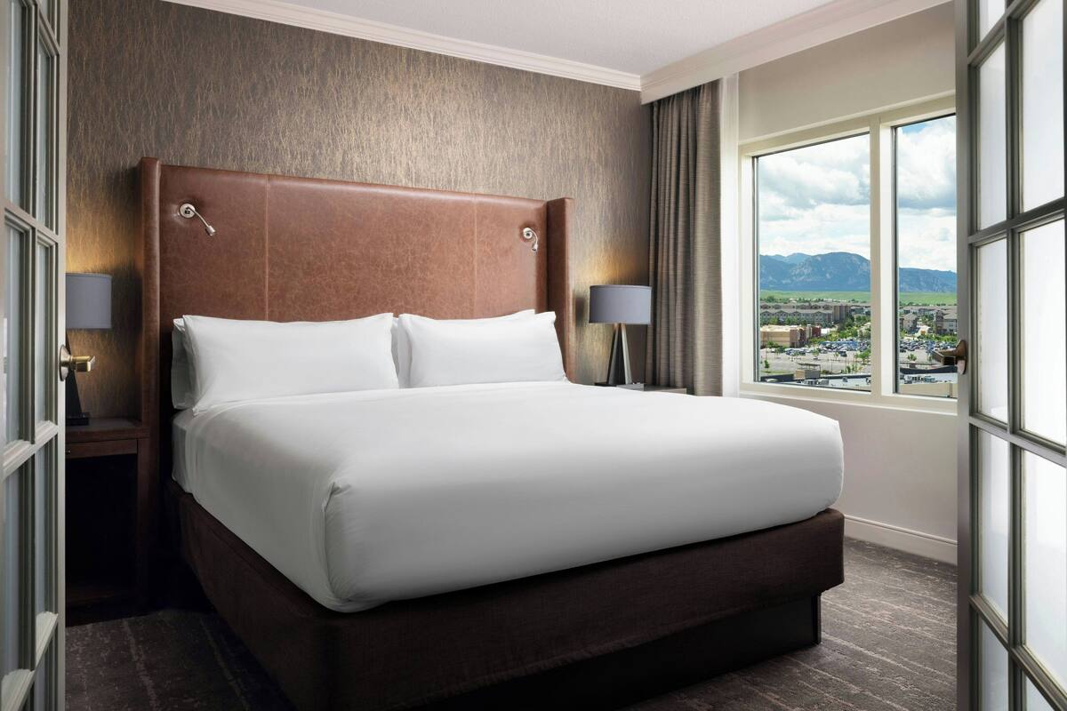 A hotel room with a large bed, two bedside tables with lamps, a view of a cityscape through large windows, and a textured wall.