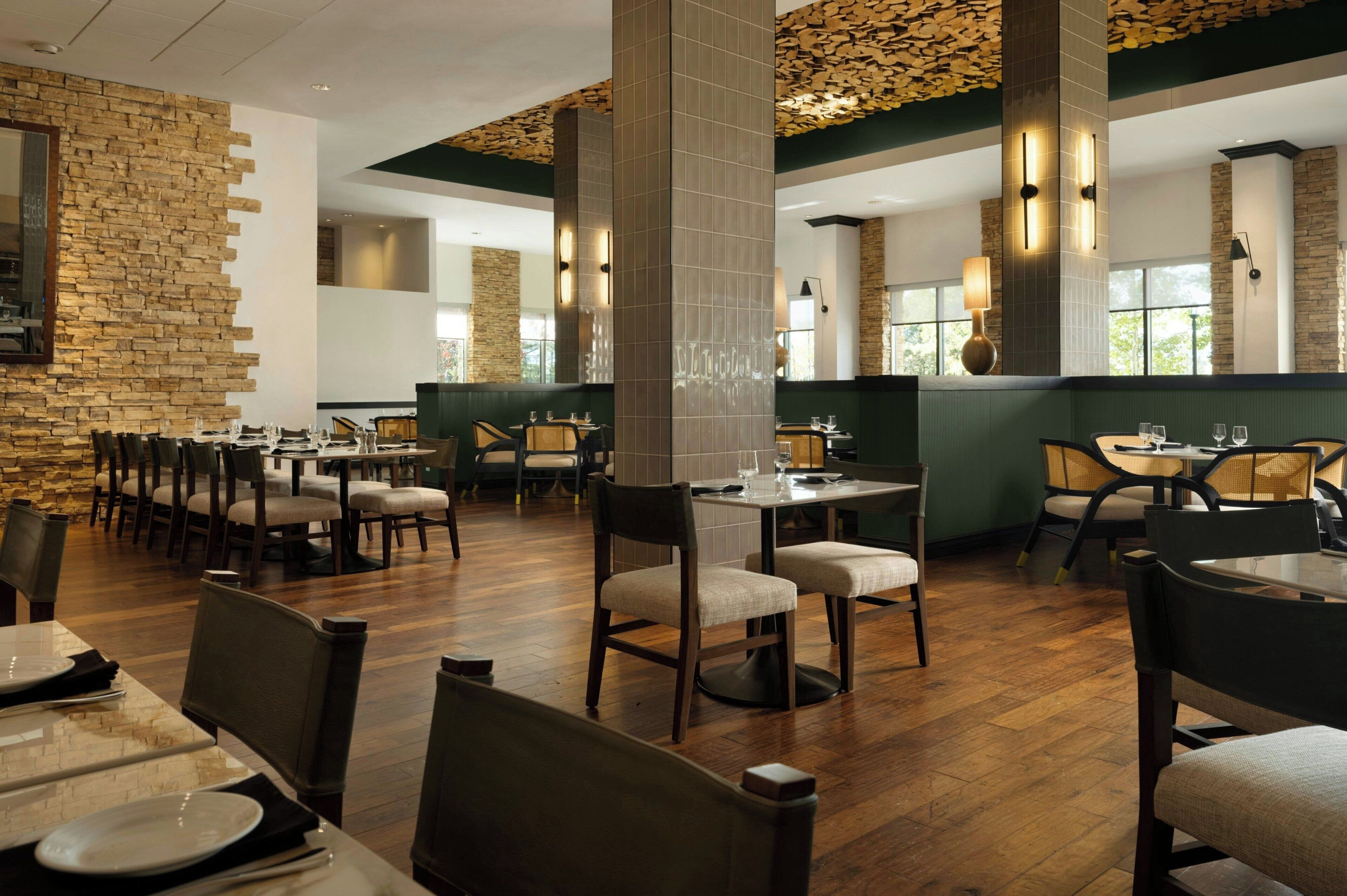 A restaurant with wooden flooring, stone walls, and modern lighting fixtures. Tables and chairs are arranged for dining.