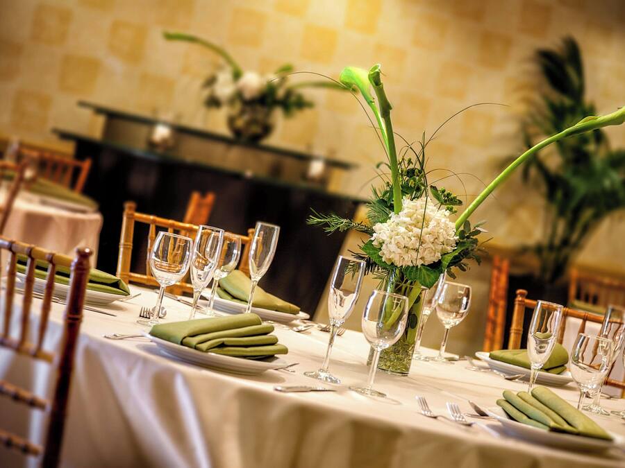 Una mesa de comedor dispuesta con servilletas verdes, platos blancos y copas de cristal, rodeada de sillas de madera.