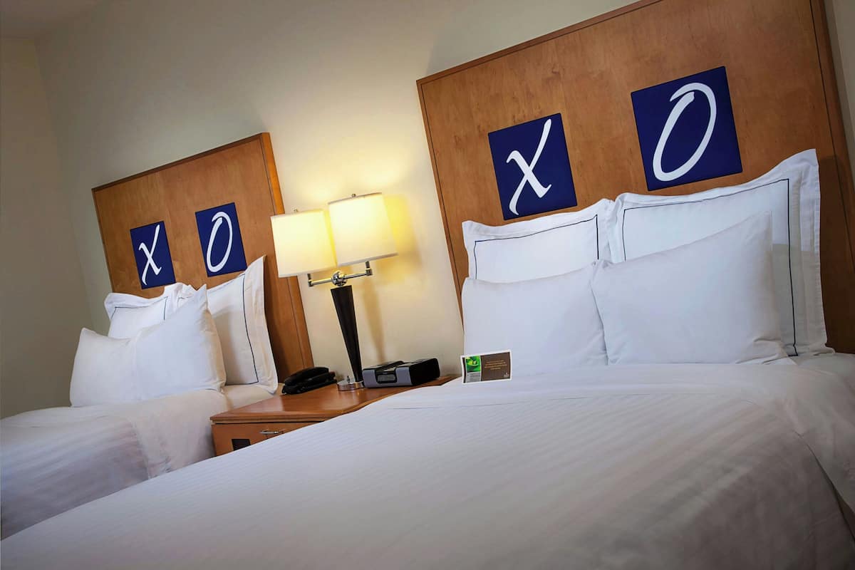 A hotel room with two beds, each with a headboard displaying 'X' and 'O' signs, a bedside table with a lamp, and a small card on the table.