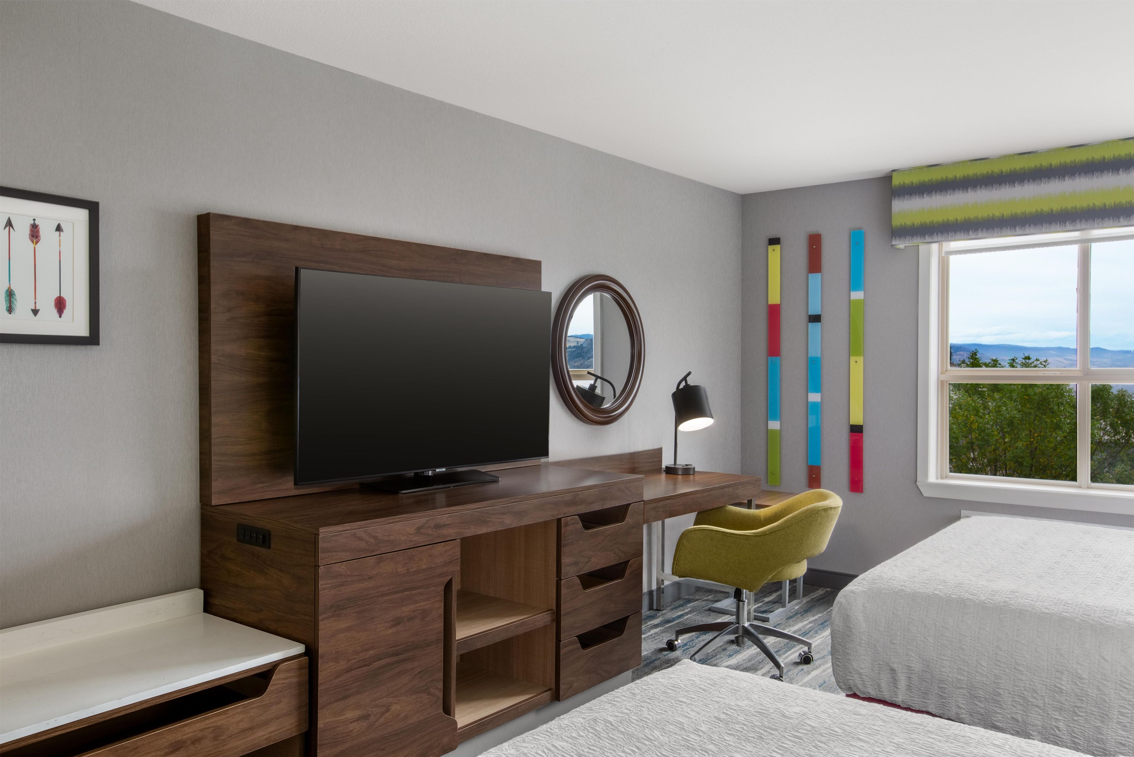 A modern hotel room with a wooden desk, a flat-screen TV, a mirror, a desk lamp, a yellow chair, a bed, and a window with a view.