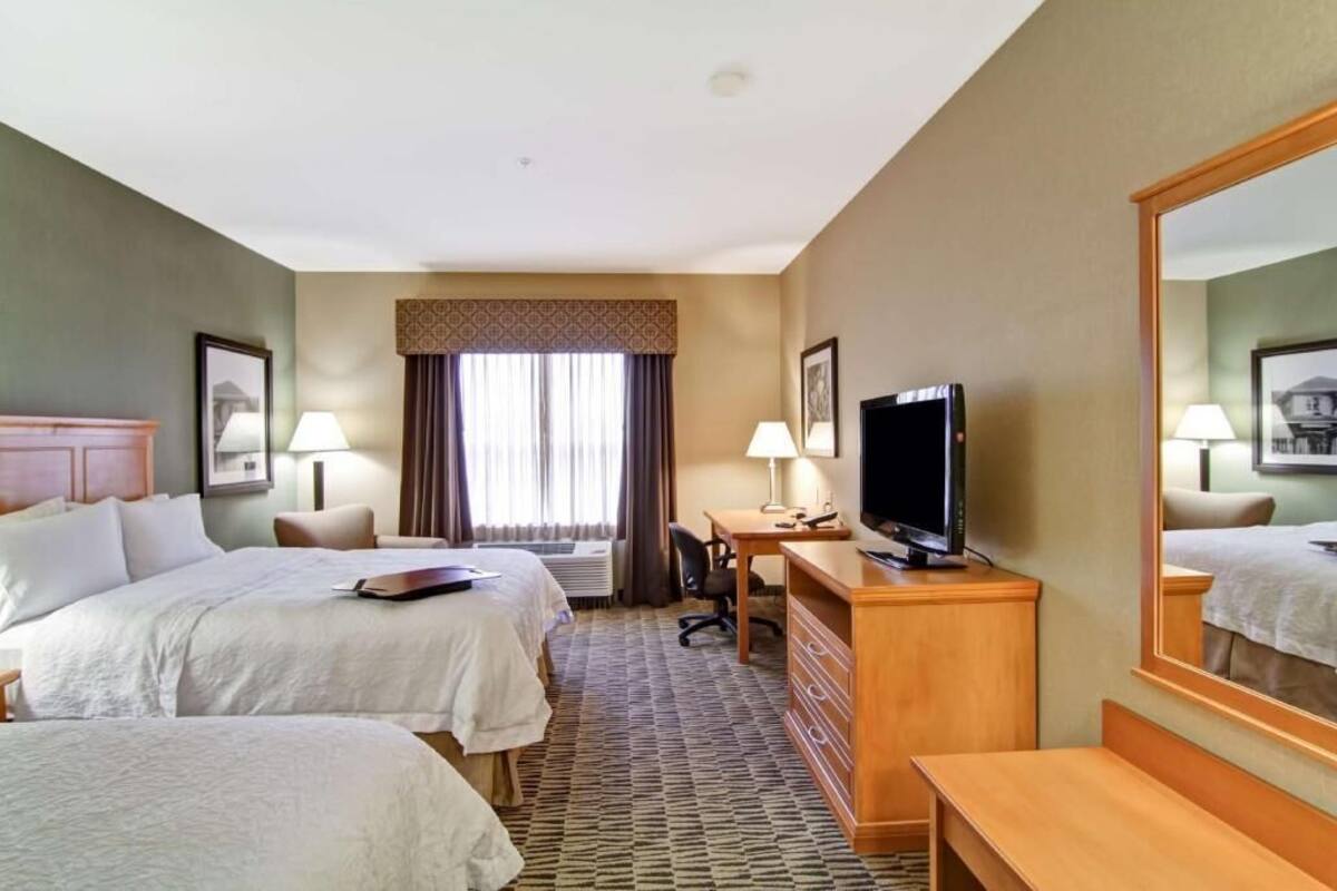 A hotel room with two beds, a desk with a television, a mirror, and a window with curtains.
