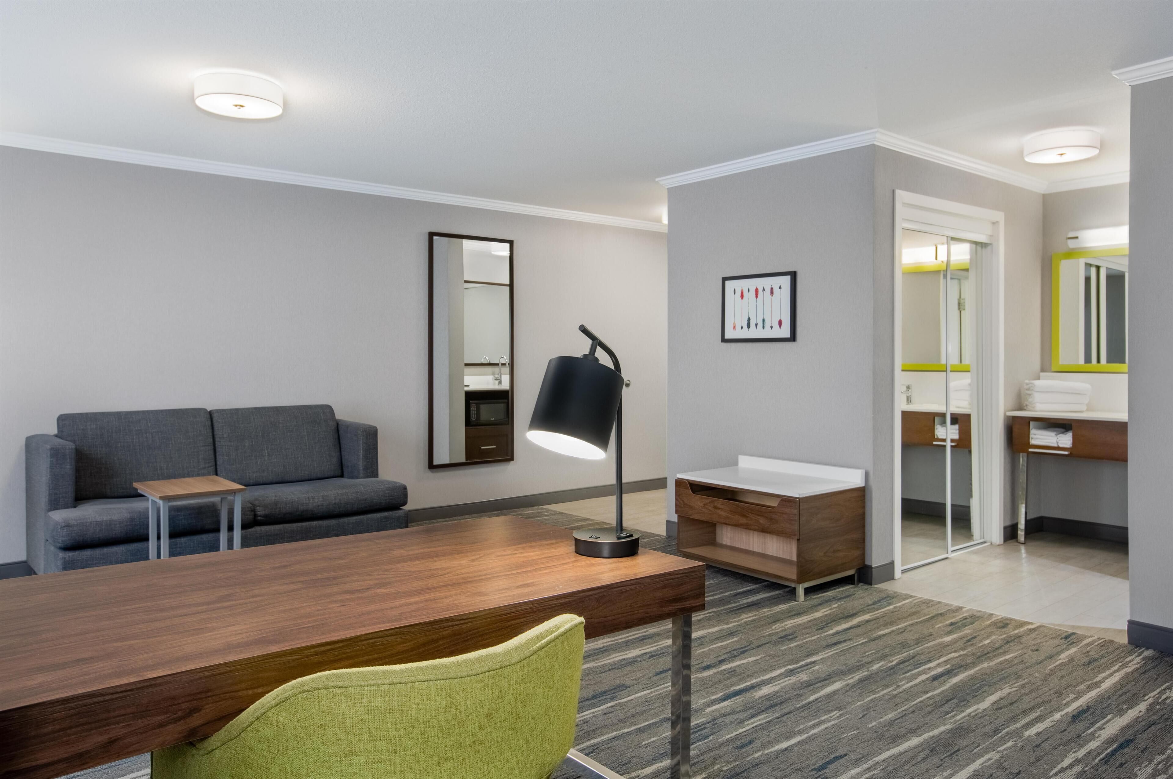 Une chambre d’hôtel moderne avec un bureau en bois, une chaise verte, un canapé gris, un miroir et une salle de bain dotée d’une porte en verre.
