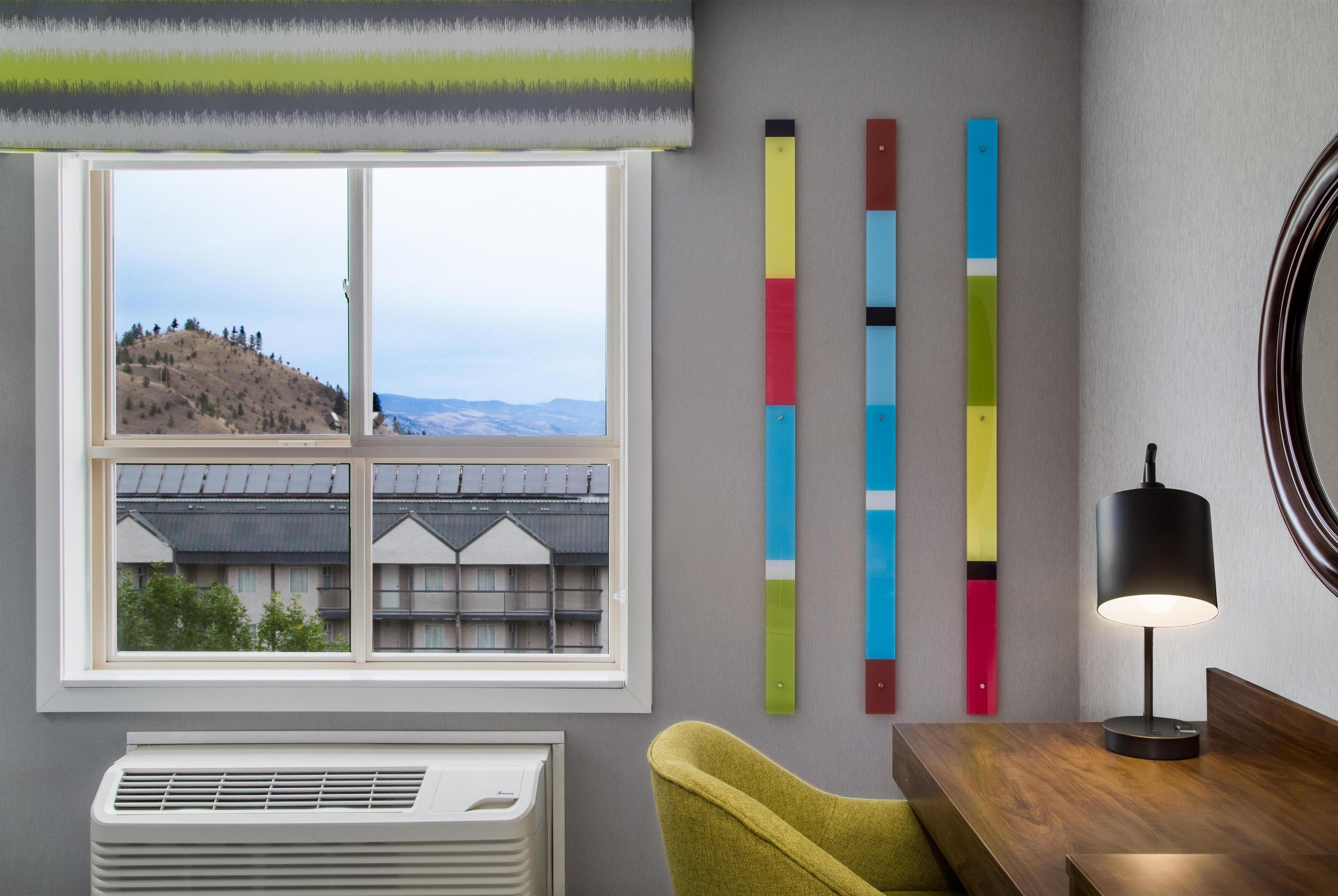 A room with a large window, a radiator, a green chair, a wooden table, and colorful vertical strips on the wall.
